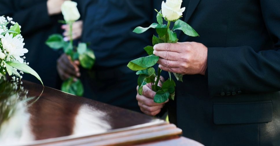 Pessoas em trajes formais pretos segurando rosas brancas diante de urnas mortuárias no contexto de costumes funerários pelo mundo.